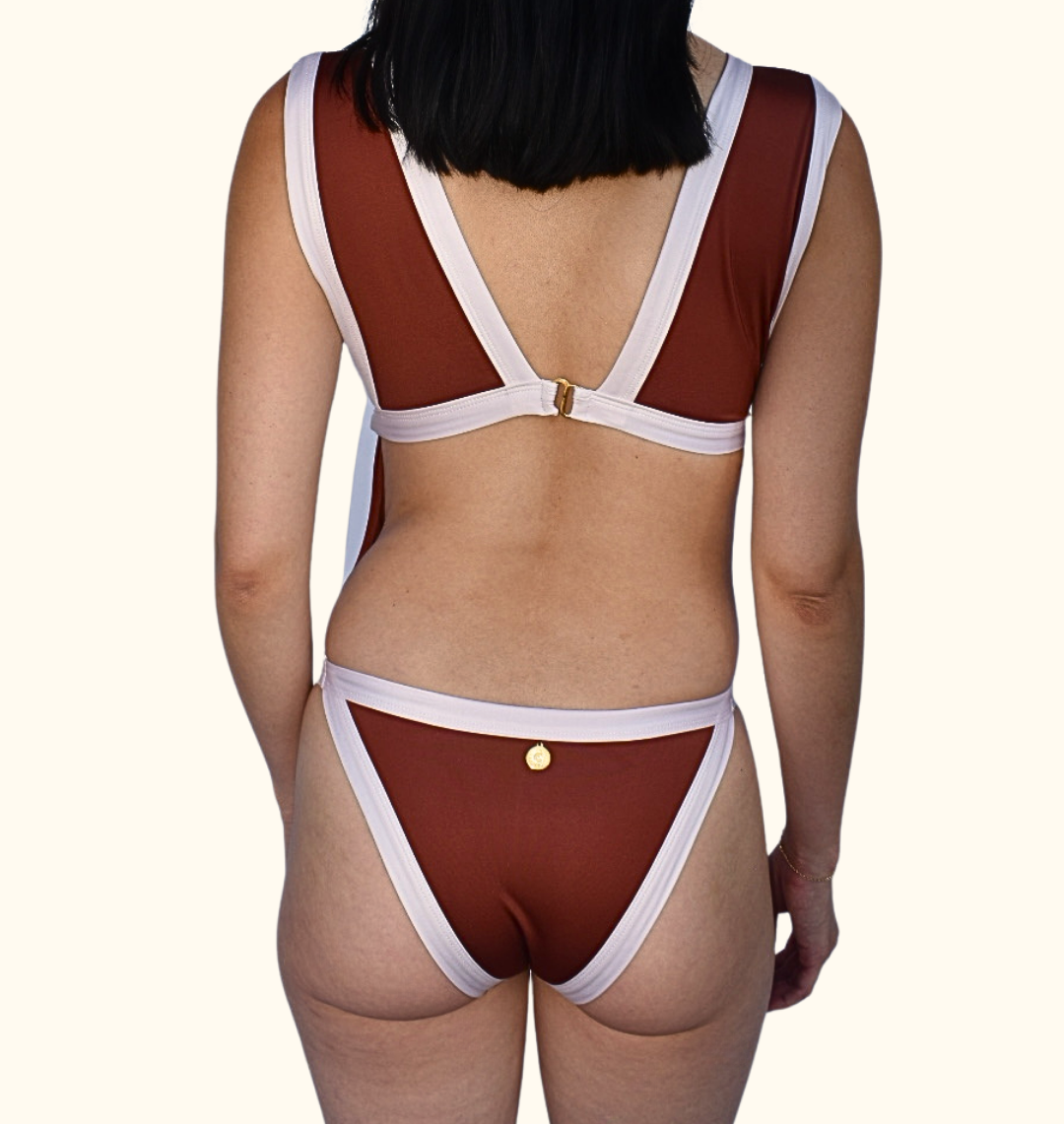 Rear view of a model wearing a two-piece bikini with a rust red body and contrasting white straps and trim, featuring small gold button details on the back and front.