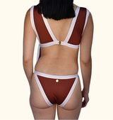 Rear view of a model wearing a two-piece bikini with a rust red body and contrasting white straps and trim, featuring small gold button details on the back and front.