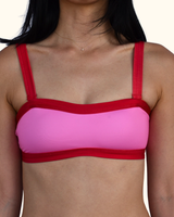 A close-up of a woman wearing a bright pink bikini top with contrasting red straps, underband, and trim, set against a plain off-white background.