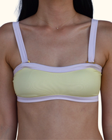 A close-up of a woman wearing a light yellow ribbed bikini top with lavender straps and gold-tone hardware, shown against a plain light background.