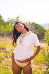 Woman in a white shirt and yellow bikini bottoms standing in a natural setting with greenery.