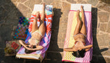Two women wearing triangle bikini sets sunbathing on colorful towels with books and a magazine.