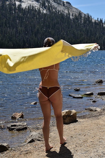 A woman stands on a rocky lakeshore facing a mountain forest, holding a large yellow towel or sarong spread across the air. She is wearing a brown string bikini.