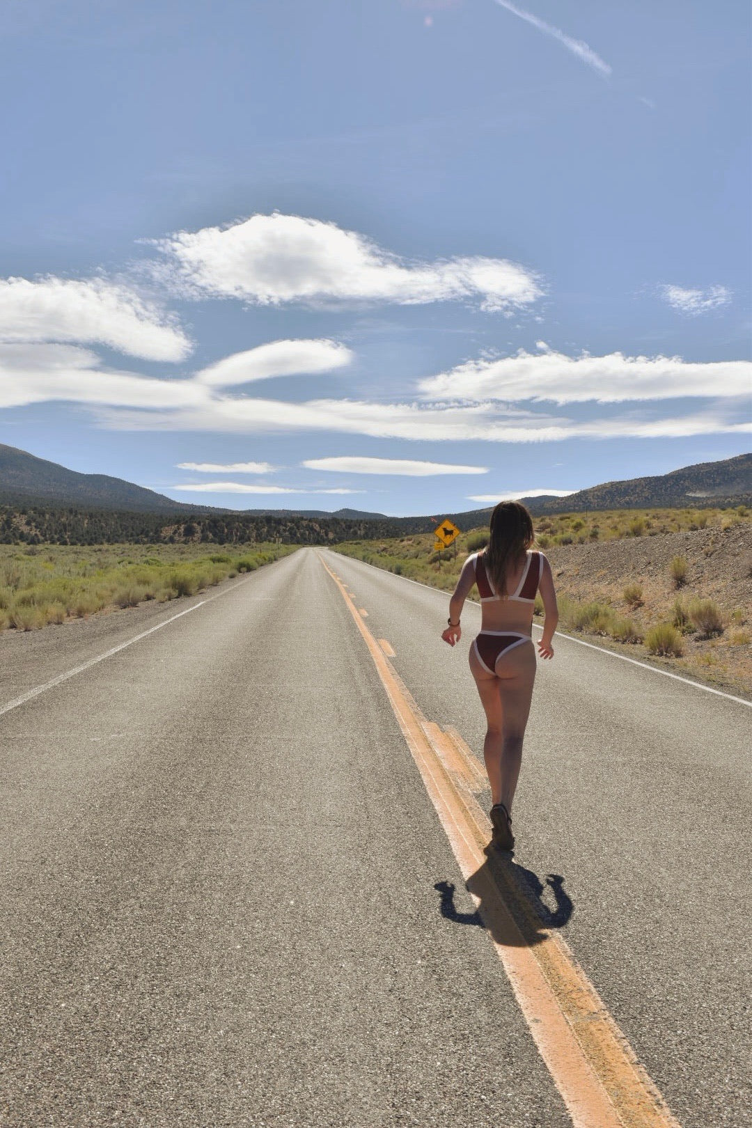A woman in a bikini walks away on a straight desert road under a blue sky with clouds.