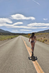 A woman in a bikini walks away on a straight desert road under a blue sky with clouds.