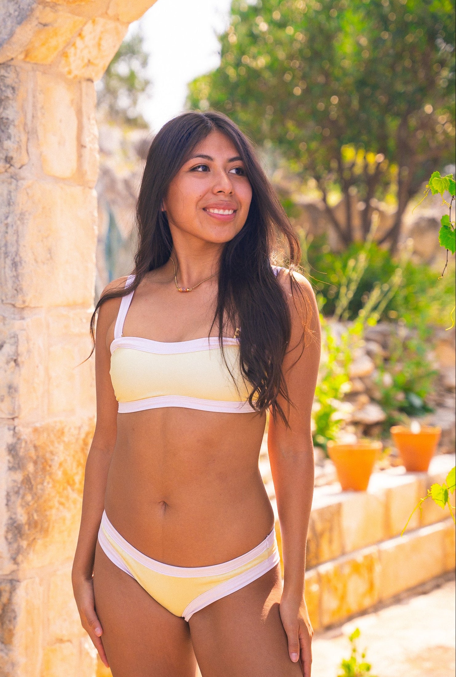 Woman in a yellow bikini standing outdoors with greenery and stone wall in the background