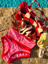 Colorful swimwear including a pink bikini bottom and red and white striped swimsuit on a stone ledge with a pool in the background.