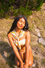 Woman wearing yellow strapless bikini set sitting outdoors with a floral scarf around her neck, surrounded by greenery.