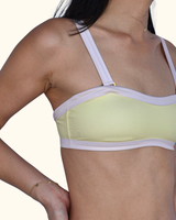 A close-up of a woman wearing a pale yellow underwire bra with light lavender straps and trim, set against a solid cream background.