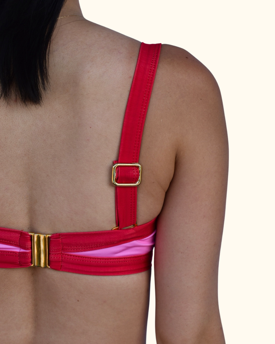 Close-up back view of a person wearing a red bikini top with gold-tone metal hardware and a pink underlayer visible at the sides, against a plain light beige background.