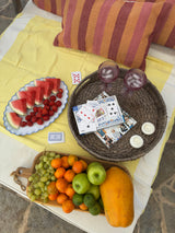 Fruits, cards, and a woven basket on a yellow cloth with a striped pillow.