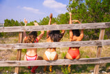 Three women in swimsuits sitting behind a wooden fence with a natural landscape in the background.