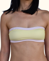 A close-up of a woman wearing a pale yellow strapless bandeau bikini top with light lavender trim, set against a plain off-white background.