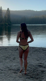 A woman stands on a sandy lakeshore at sunrise, viewed from behind, wearing a yellow bikini with black trim. Trees and a calm lake are visible in the background under a clear blue sky.