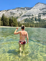 A woman stands in shallow, clear green lake water with a mountain, pine trees, and blue sky behind her, wearing a white bikini top and dark brown bikini bottoms.