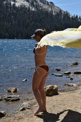 A woman stands on a rocky lakeshore wearing a dark brown string bikini and a gray baseball cap, holding a large pale yellow beach towel or blanket in front of her. A blue lake, dense evergreen forest, and rocky mountains are visible in the background.